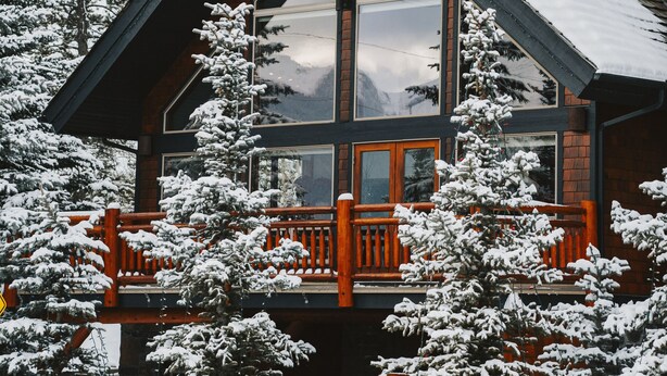 Une cabane dotée d’une grande baie vitrée, d’une terrasse en bois et d’arbres couverts de neige.