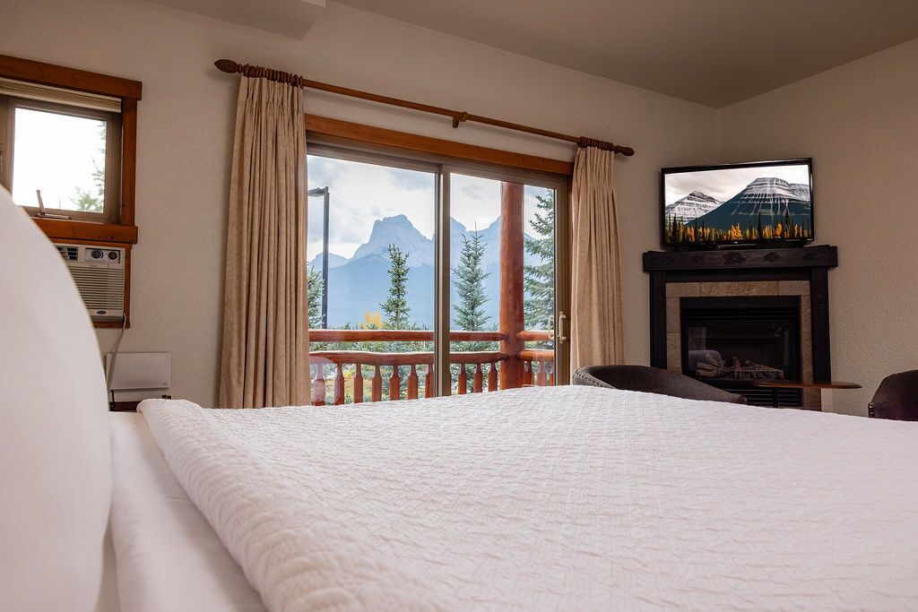 A hotel room with a large bed, a television mounted above a fireplace, and a balcony with a mountain view.