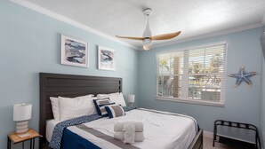 A neatly made bed with a blue and white bedspread, a wooden nightstand with a lamp, and a ceiling fan. - Clearwater Beach Suites #103 | Ground floor + Heated Pool + Walk to the Beach! (Clearwater Beach)