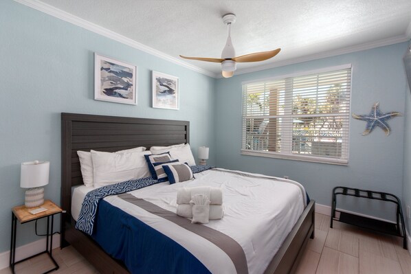 A neatly made bed with a blue and white bedspread, a wooden nightstand with a lamp, and a ceiling fan. - Clearwater Beach Suites #103 | Ground floor + Heated Pool + Walk to the Beach! (Clearwater Beach)