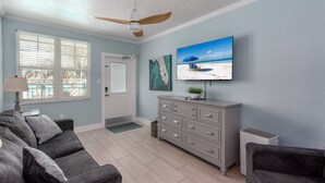 A living room with a gray sofa, a gray cabinet, a ceiling fan, a TV on the wall, and a window with blinds. - Clearwater Beach Suites #103 | Ground floor + Heated Pool + Walk to the Beach! (Clearwater Beach)
