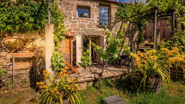 Una casa de piedra con un letrero que dice 'CASA DA EVA', un pequeño porche y un jardín con flores amarillas y enredaderas.