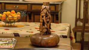 Una lámpara de mesa con base de madera y pantalla decorativa, un cuenco de naranjas y un piano al fondo.