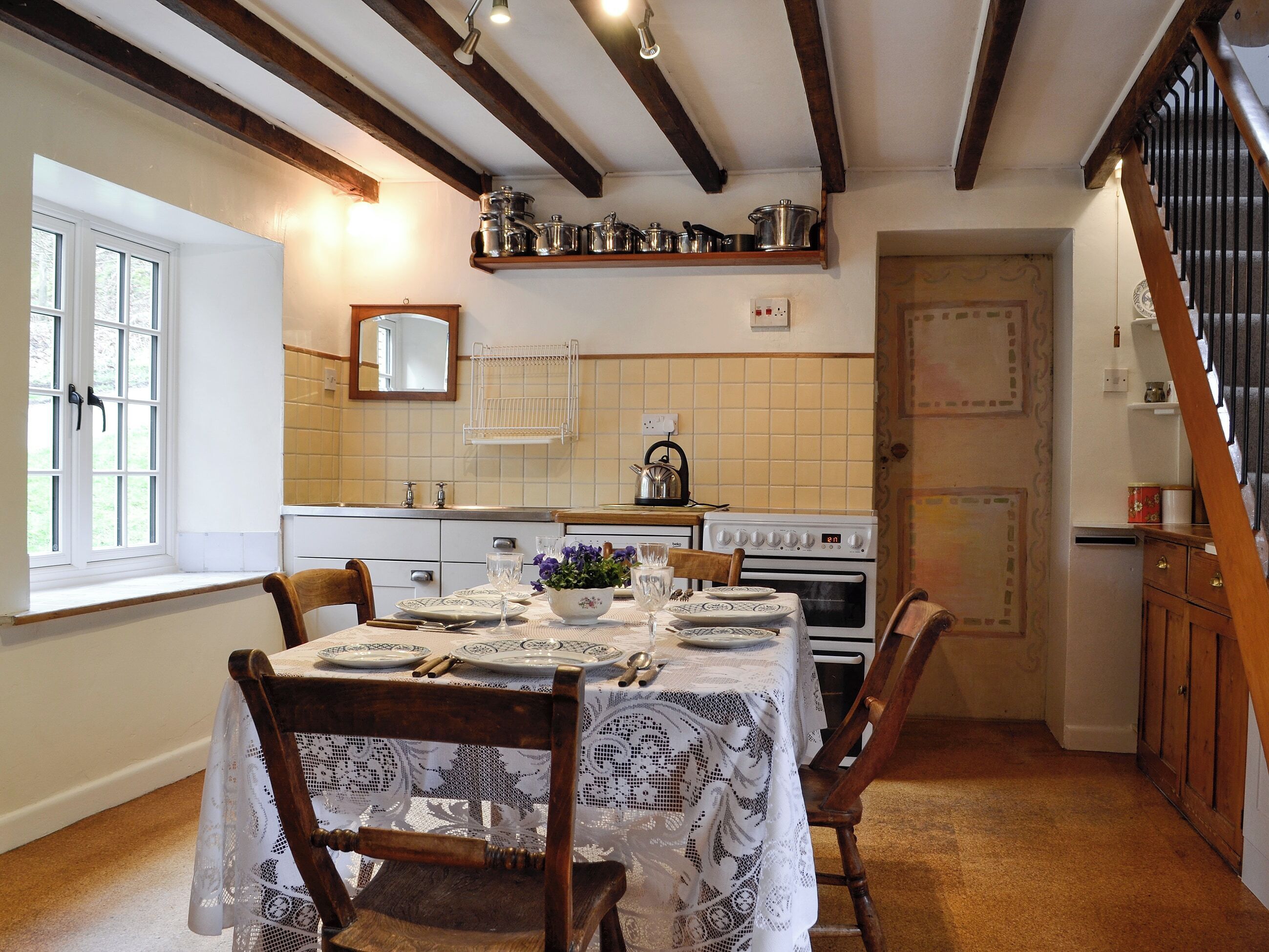 A dining room with a table set for four, a wooden chair, a kitchen with a stove, and a window with a view of the outdoors.