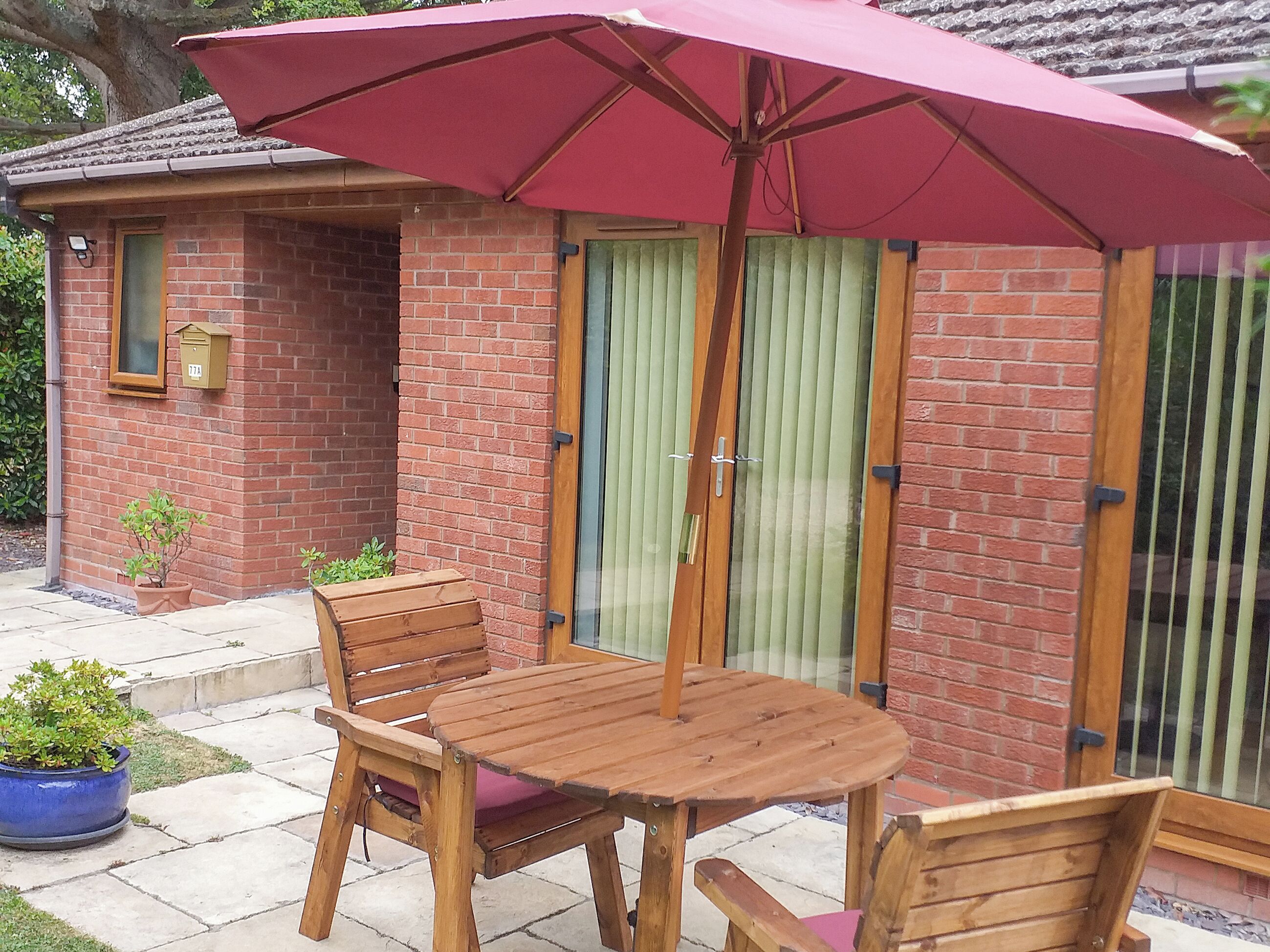 Eine Terrasse mit Holztisch und -stühlen, einem roten Sonnenschirm und einem Backsteinhaus mit grüner Tür.