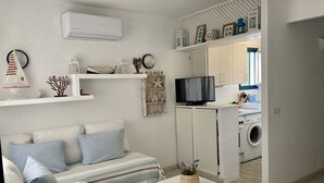 A modern living room with a sofa, a small table, and a TV. There is a kitchen area with a washing machine visible.
