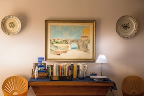 A room with a painting of a coastal scene, a bookshelf with books, a lamp, and two chairs.