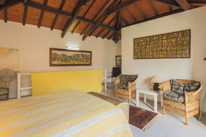 A room with a bed, two wicker chairs, a small table, a fan, and a large artwork on the wall.