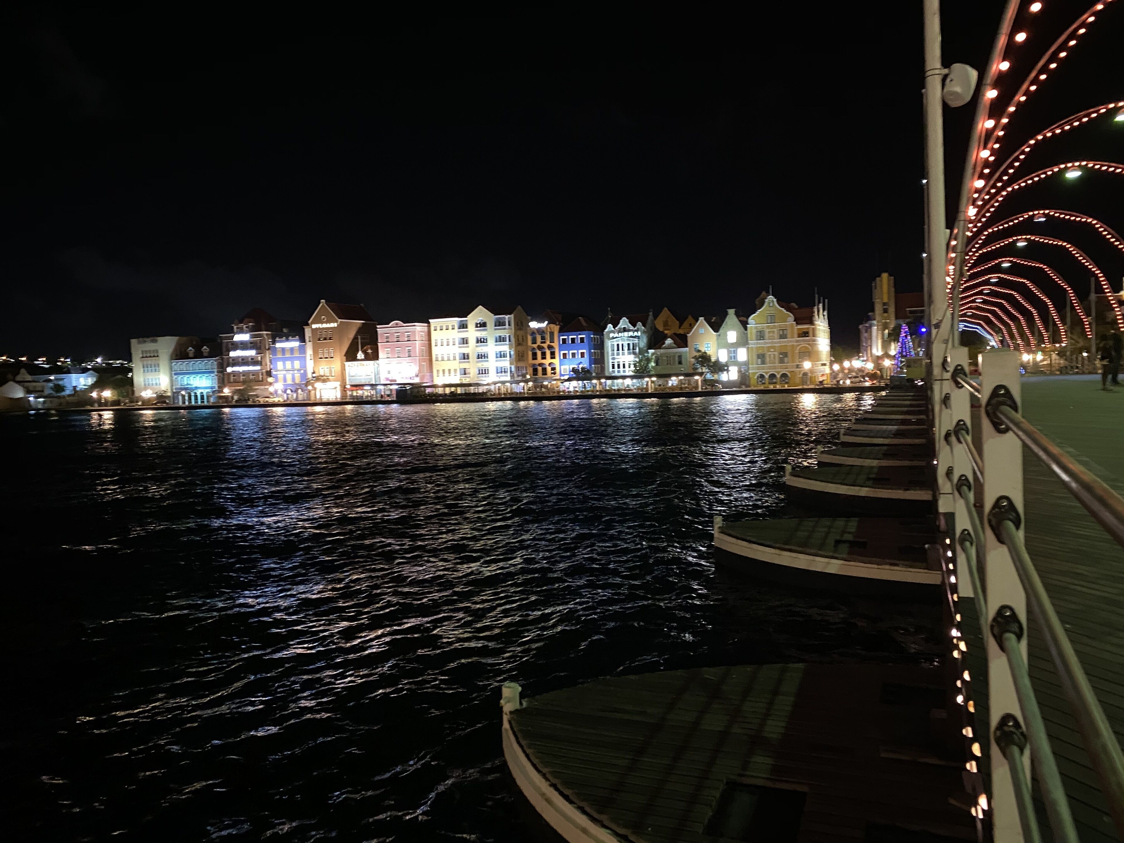 A waterfront view with illuminated buildings and a bridge railing.