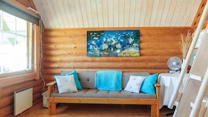 Interior acolhedor de uma cabana com soalho de madeira, um sofá com almofadas azuis e brancas, um quadro de peixes na parede e um ventilador sobre uma mesa.