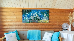 Interior de cabana de madeira com teto de madeira, sofá xadrez com almofadas azuis e brancas, e um quadro de peixes pendurado na parede.