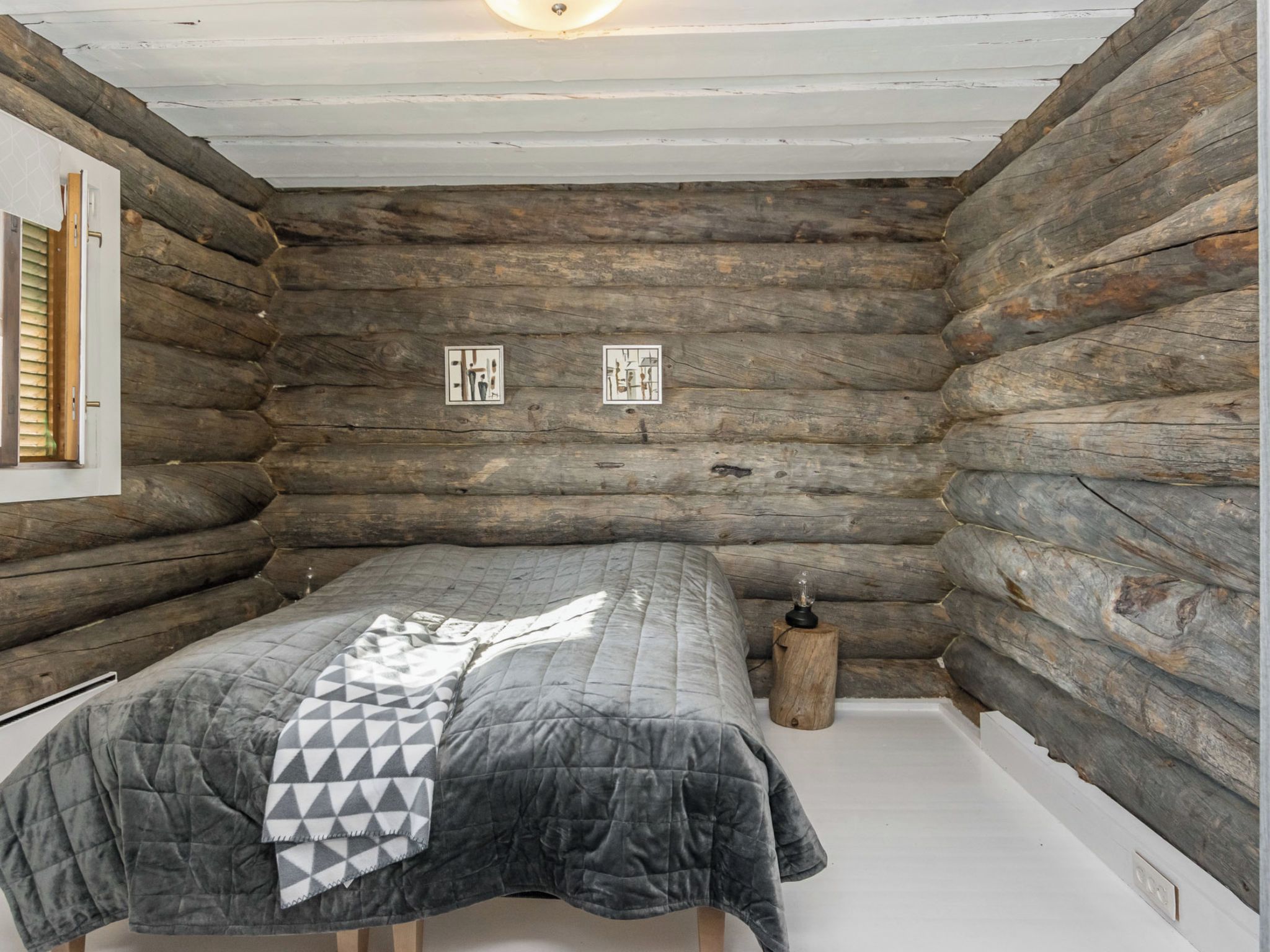 Une chambre de style rustique, avec un lit, une fenêtre équipée de stores et une petite table avec une lampe.