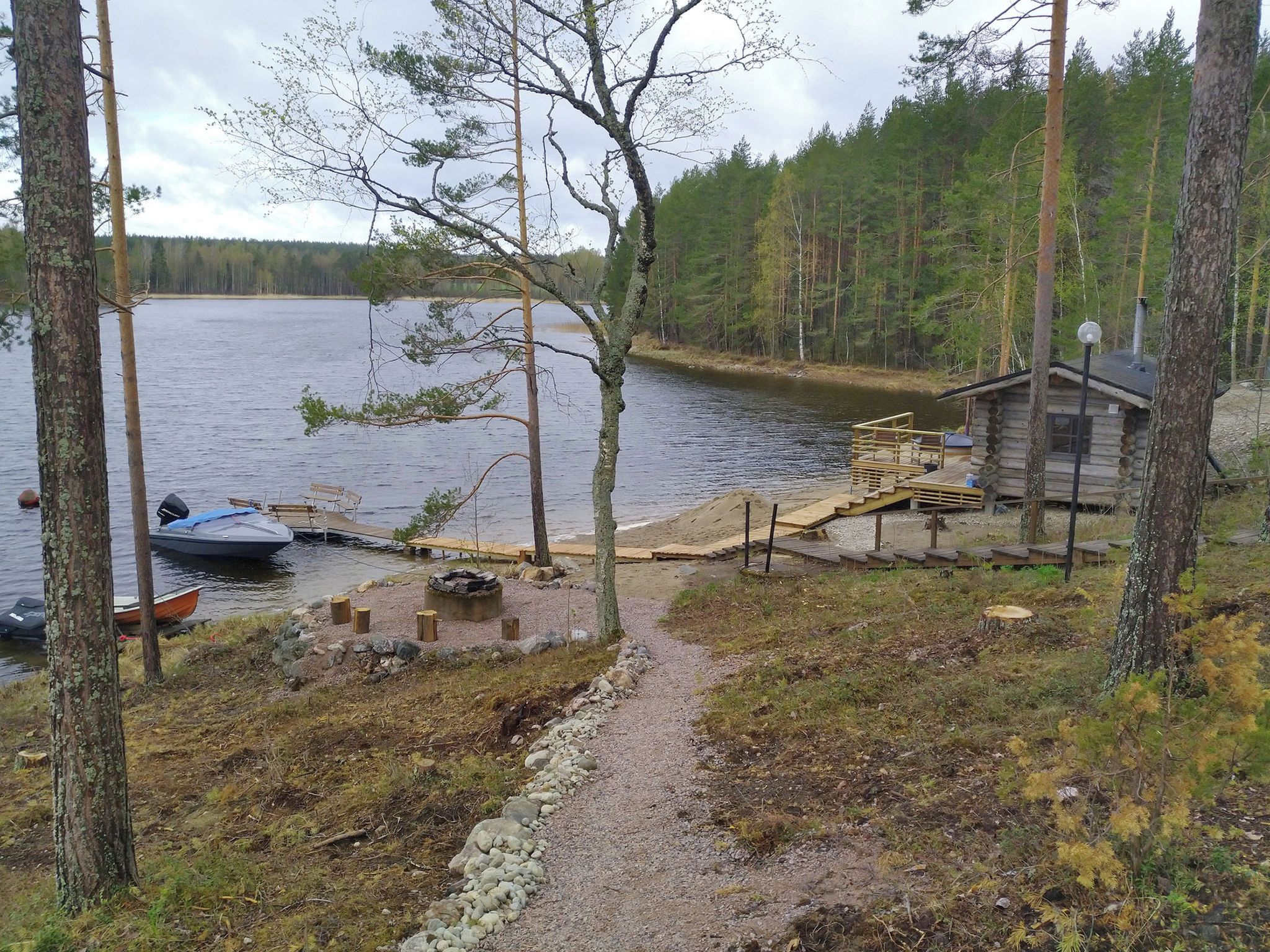 Une cabane au bord d’un lac, avec un quai, un bateau et un foyer extérieur.