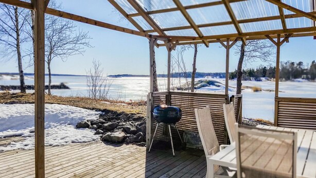 Une terrasse en bois avec un toit de verre donnant sur un lac gelé, un barbecue et une table avec des chaises.