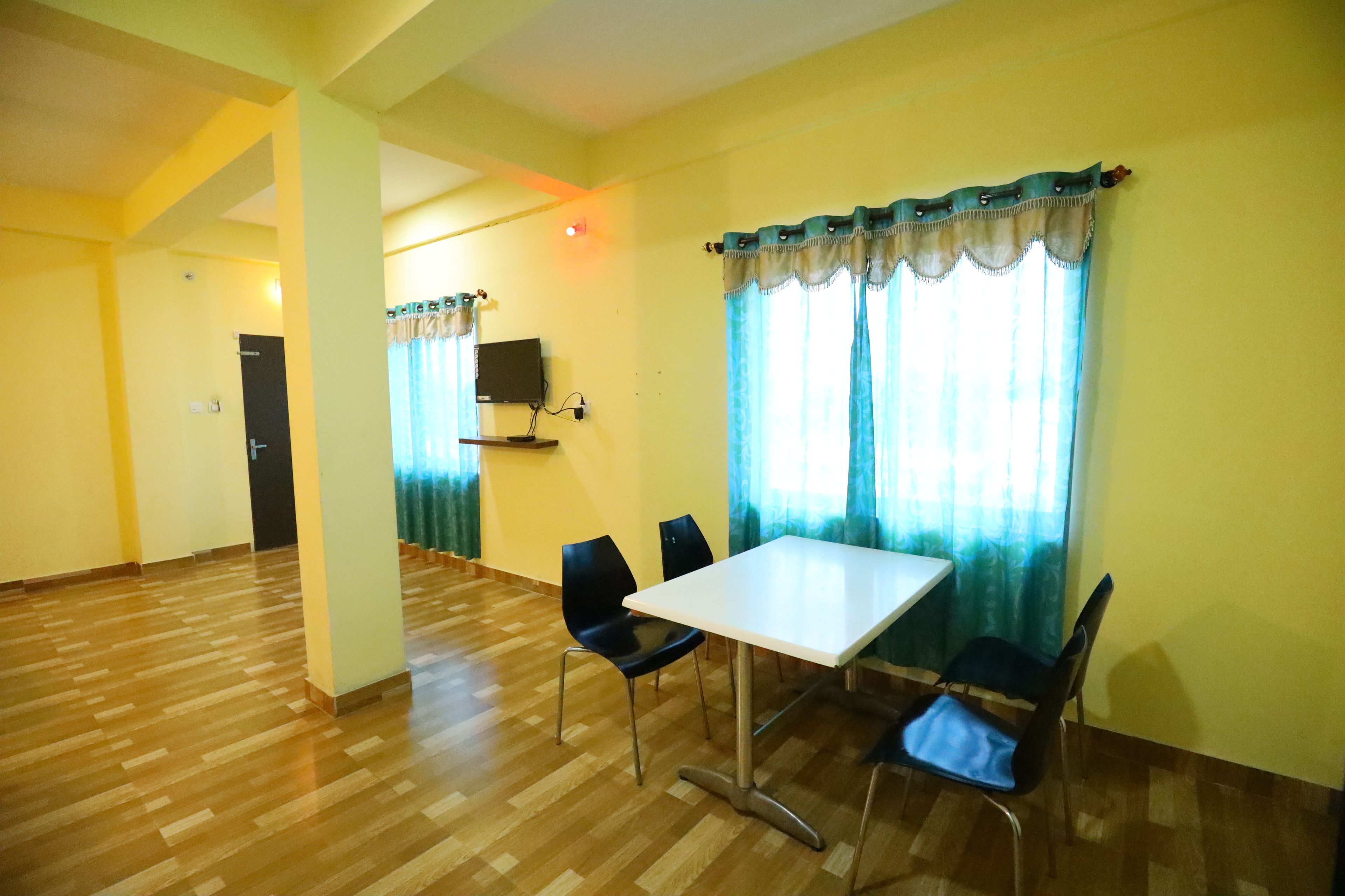 Una habitación con piso de madera, una mesa blanca y cuatro sillas negras. Se ve una puerta al fondo y una ventana con cortinas.