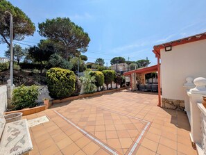 A courtyard with a tiled floor, surrounded by various plants and trees, and a building with a red roof.
