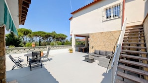 Un patio techado con mobiliario exterior, un muro de piedra y una escalera que conduce a un nivel superior.