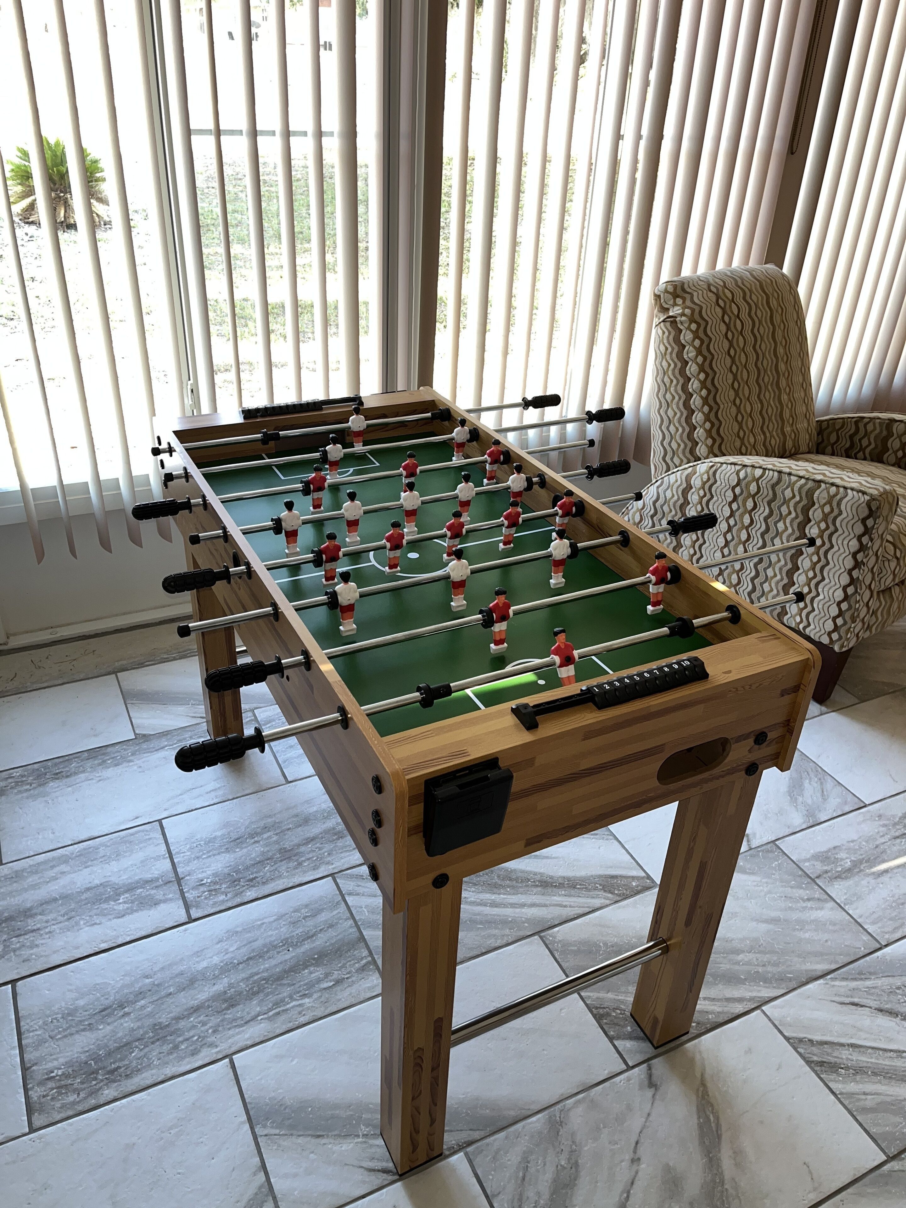 A wooden foosball table with a green playing surface, positioned in a room with tiled flooring and a patterned armchair.