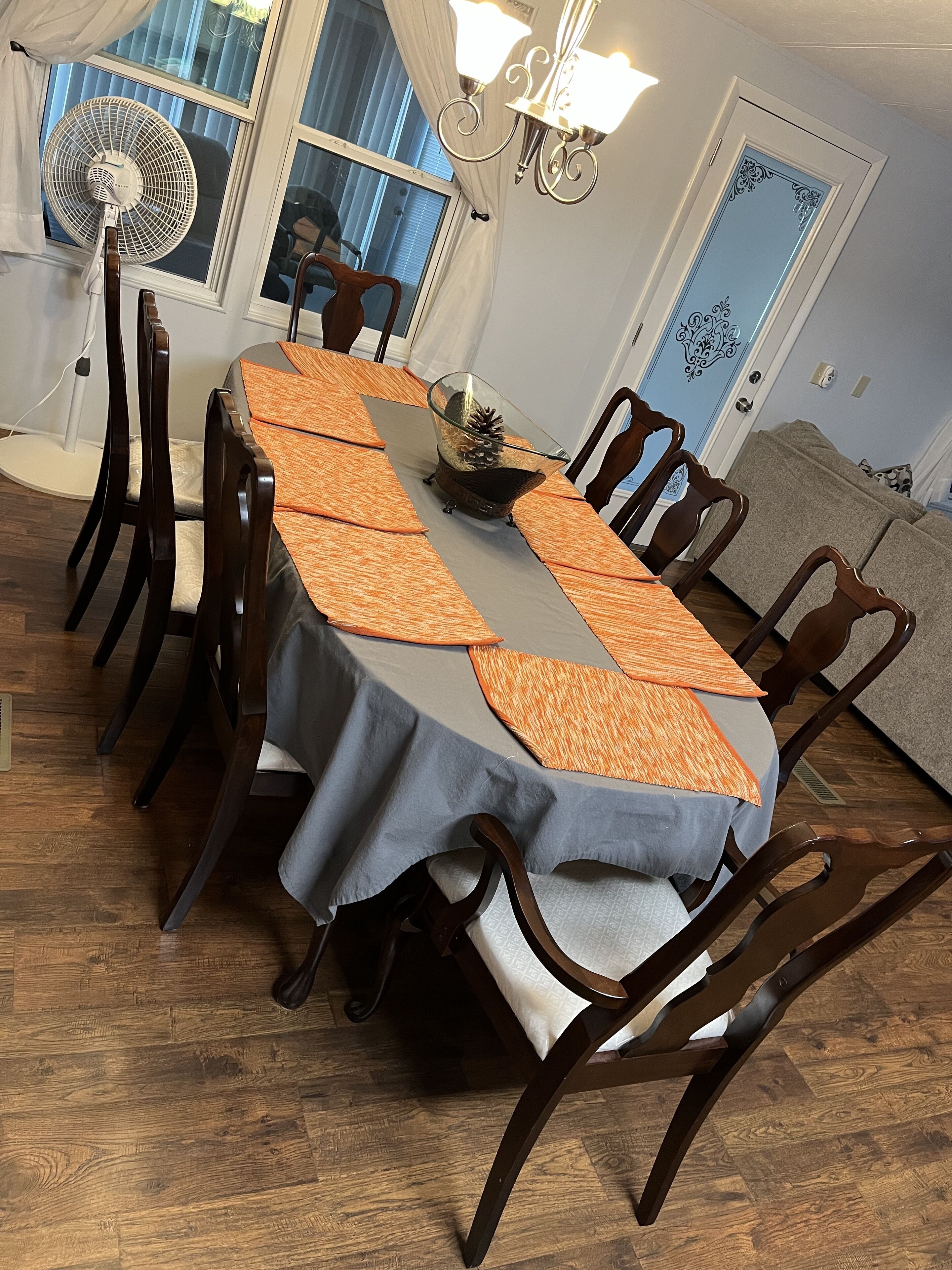 A dining area with a table set for six, surrounded by wooden chairs. A fan is on the left, and a chandelier hangs above the table.