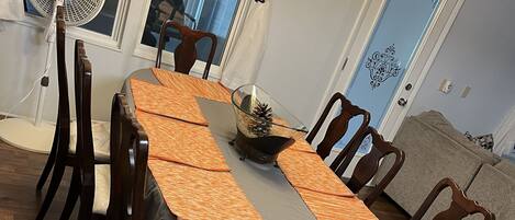 A dining area with a table set for six, surrounded by wooden chairs. A fan is on the left, and a chandelier hangs above the table.