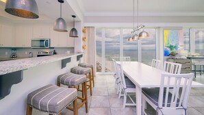 Una cocina moderna con armarios blancos, una isla grande y taburetes de bar. Hay un comedor con una mesa y sillas blancas, y una vista al océano a través de ventanas amplias.