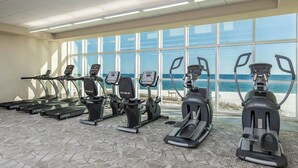A gym with various exercise machines, large windows, and a view of the beach.