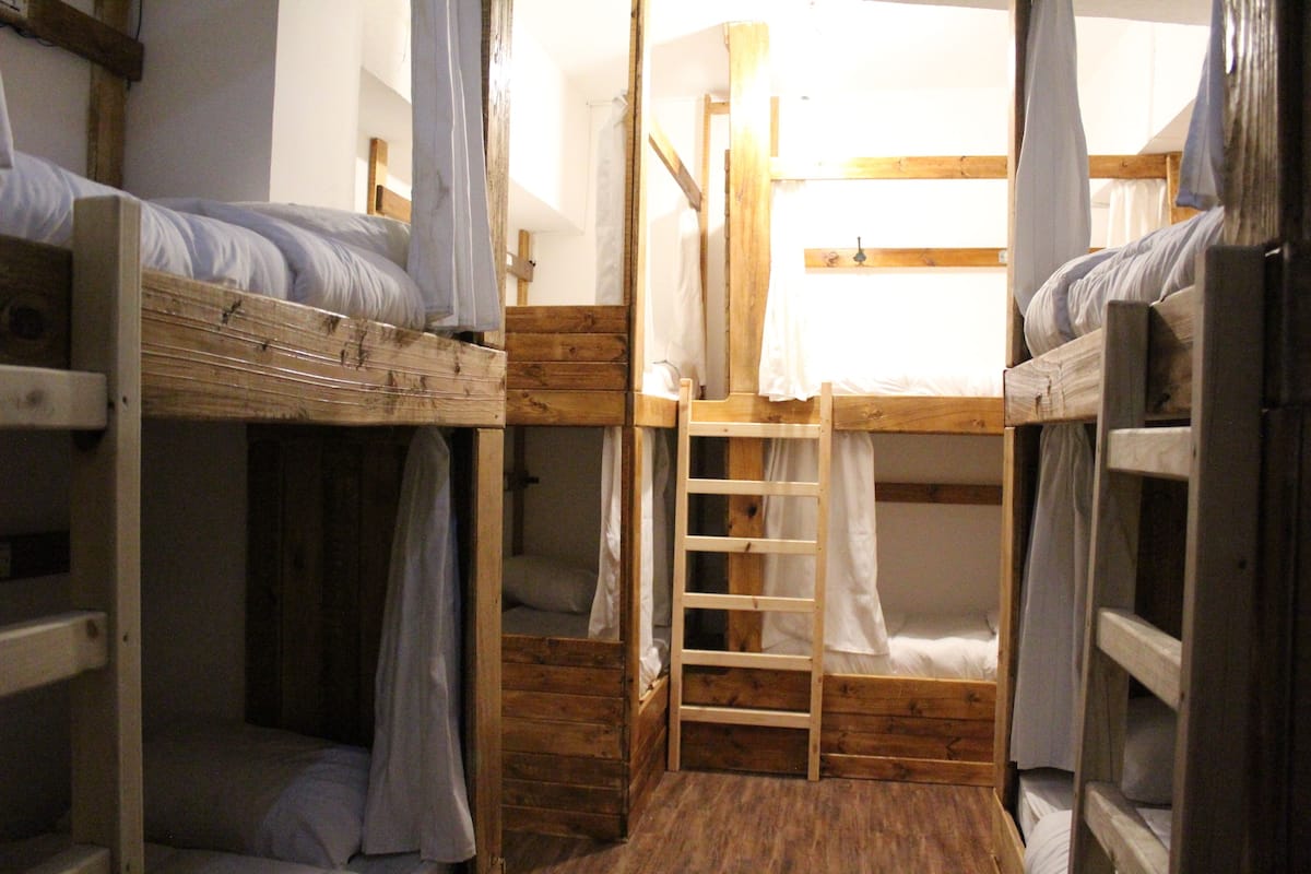 A dormitory room with bunk beds, a wooden ladder, and a window with curtains.