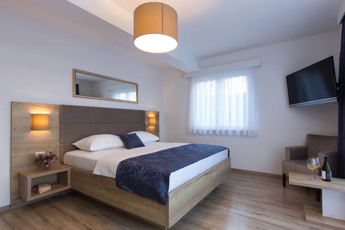 A modern hotel room with a wooden bed, bedside lamps, a flat-screen TV, a small table with a bottle and glass, and a window with curtains.