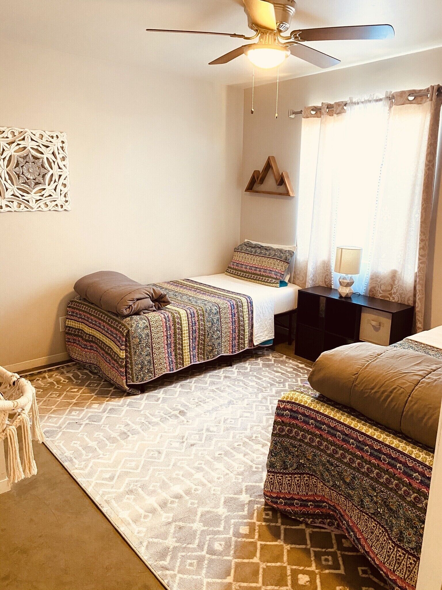 A bedroom with two beds, a ceiling fan, a decorative wall piece, and a patterned rug.