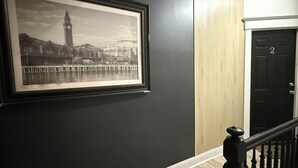Un couloir orné d’une photographie en noir et blanc encadrée représentant un paysage urbain, une porte en bois et un escalier avec une rampe noire.
