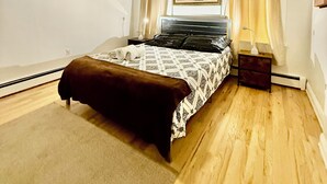 Une chambre à coucher comprenant un lit, un ventilateur de plafond, une table de chevet, une lampe, une fenêtre avec des rideaux et un élément de décoration murale.