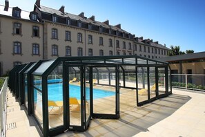 Una piscina cubierta al aire libre con paredes de vidrio, rodeada por una terraza de baldosas y una hilera de edificios al fondo.