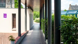 A covered walkway with columns, a wooden floor, and a building with a stone facade. - Proche du Centre | Piscine Extérieure Chauffée (Dinan)
