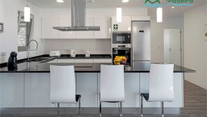 A modern kitchen with a central island, stainless steel appliances, and white bar stools. - Beautiful Modern Villa in Calheta – CalhetaScape (Estreito da Calheta)