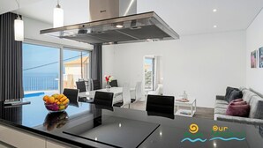 A modern kitchen with a large island, a dining area, and a living space with a view. - Beautiful Modern Villa in Calheta – CalhetaScape (Estreito da Calheta)
