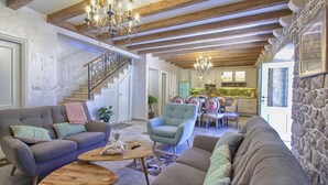 Ein geräumiges Wohnzimmer mit hohen Decken, freiliegenden Holzbalken und einem Chandelier. Eine Treppe führt zu einer weiteren Ebene, es gibt einen Essbereich mit Tisch und Stühlen sowie eine Küche im Hintergrund.