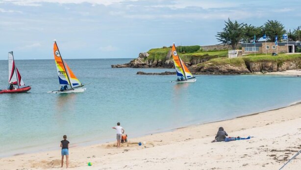 Una playa con tres veleros, una orilla arenosa y un edificio con banderas al fondo.