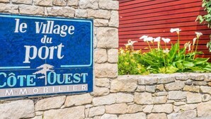 Un cartel para 'le Village du Port', con un muro de piedra y un edificio rojo al fondo.