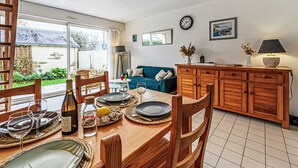 Un comedor con una mesa de madera dispuesta para cuatro personas, un sofá azul al fondo y una cocina con armarios de madera y un reloj en la pared.