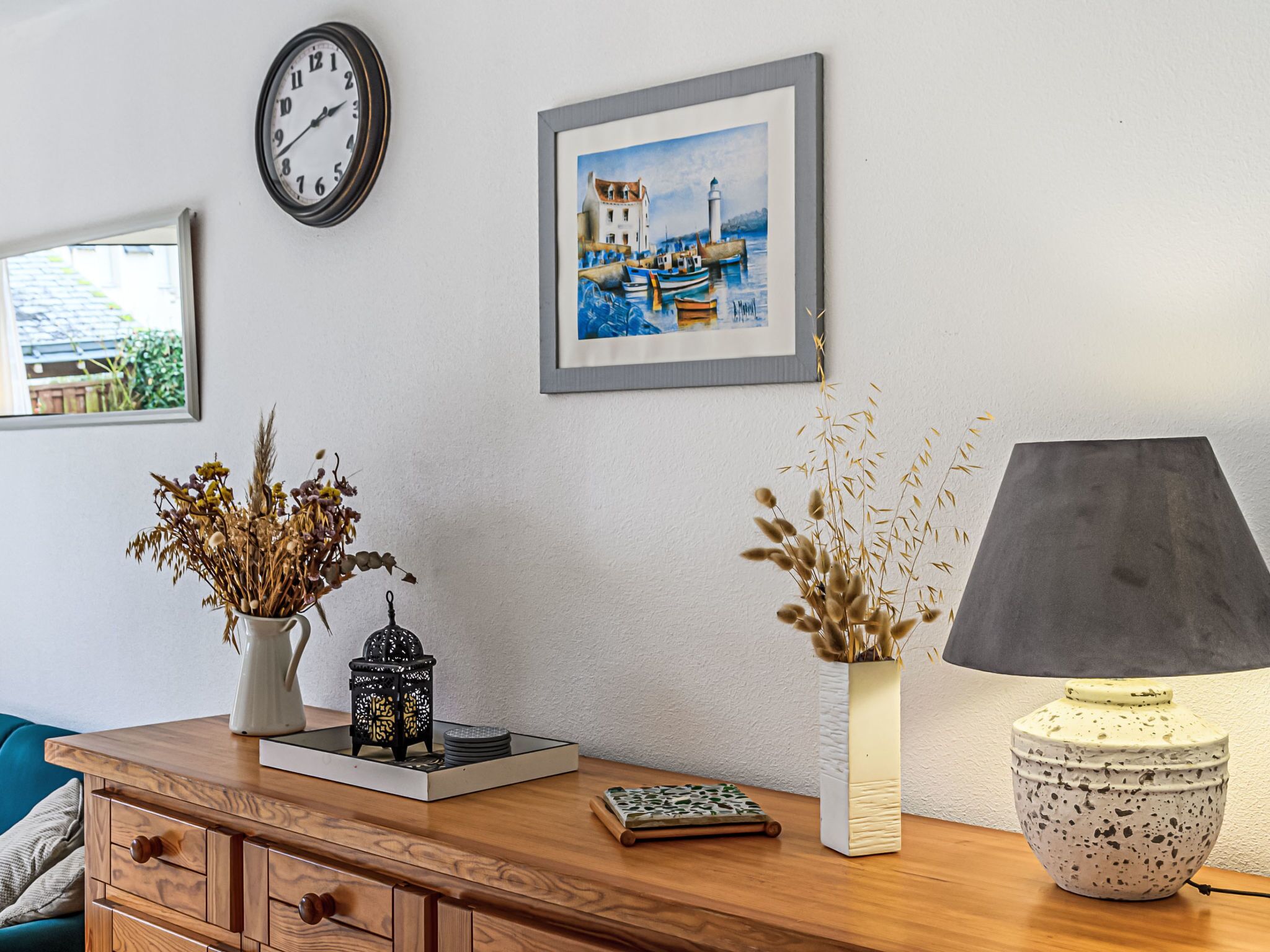 A room with a wooden dresser, a clock, a framed painting, and a table with a lamp and decorative items.