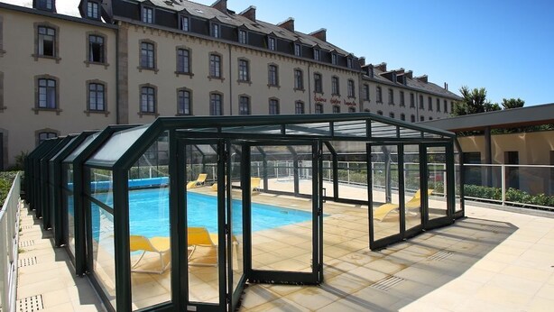 A covered outdoor swimming pool with glass walls, surrounded by a tiled deck and a row of buildings in the background.