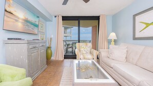 A living room with a sofa, armchair, and a coffee table with a turtle figurine. There is a ceiling fan, a painting of a beach, and a view of the balcony.