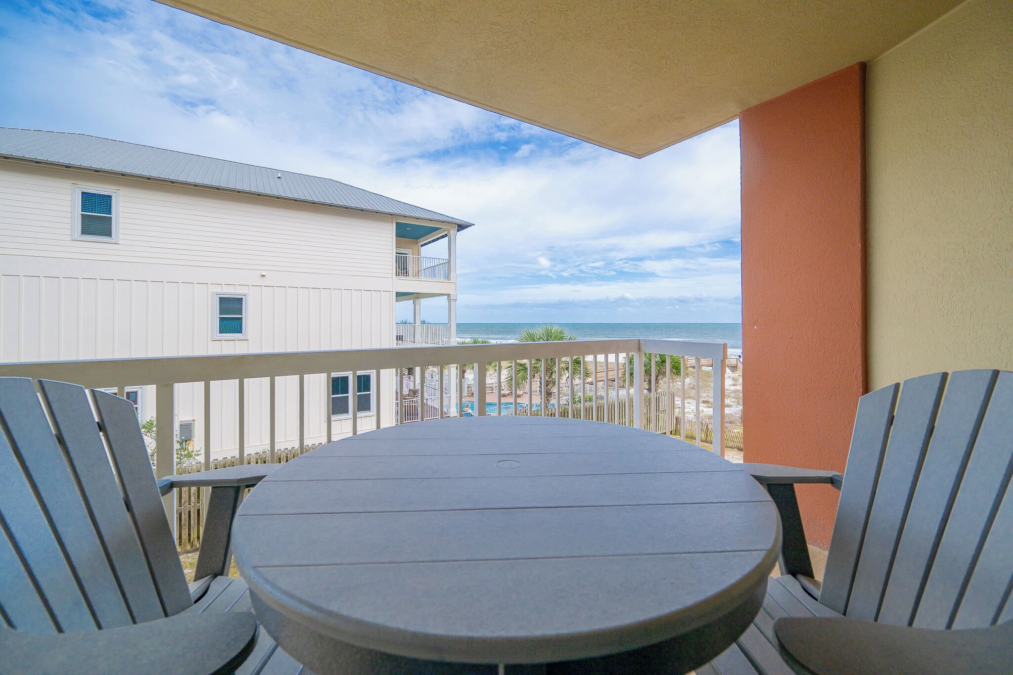 A balcony with a round table and two chairs, overlooking a beach and a building with a white exterior.