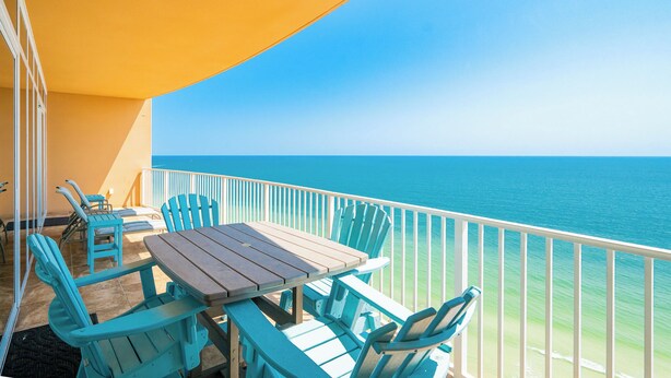 A balcony with a wooden table and turquoise chairs, overlooking a clear blue ocean. - Lazy River + Beachfront  3 Bedroom 3 Bath Phoenix Orange Beach 1905 (Orange Beach)