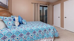 A bedroom with a large bed, beige walls, a ceiling fan, a window with curtains, and a door. - Fantastic 2BR Condo - Romar Village 622 (Orange Beach)