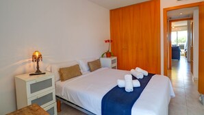 Uma cama bem arrumada com lençóis brancos e um toque de azul, uma mesa de cabeceira com luminária, uma pequena planta em vaso e uma porta de madeira que dá para outro cômodo.