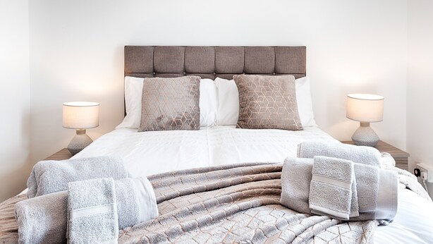 A neatly made bed with a grey and white patterned bedspread, two matching pillows, and a grey blanket. There are two bedside lamps on wooden nightstands. - Generous Home near Liberty Stadium (Swansea)