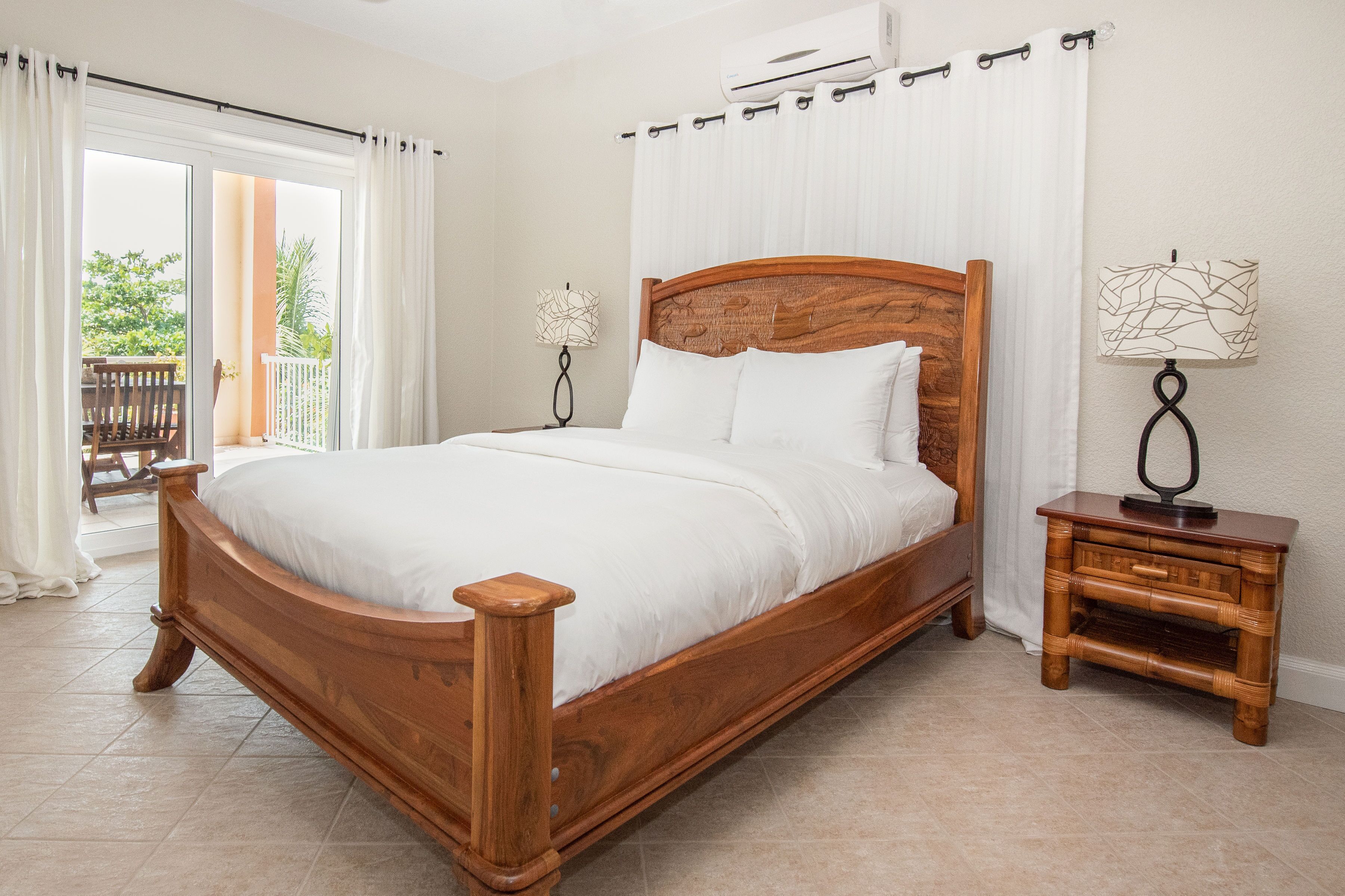 A wooden bed with white bedding, a nightstand with a lamp, and a view of a balcony with a table and chairs.