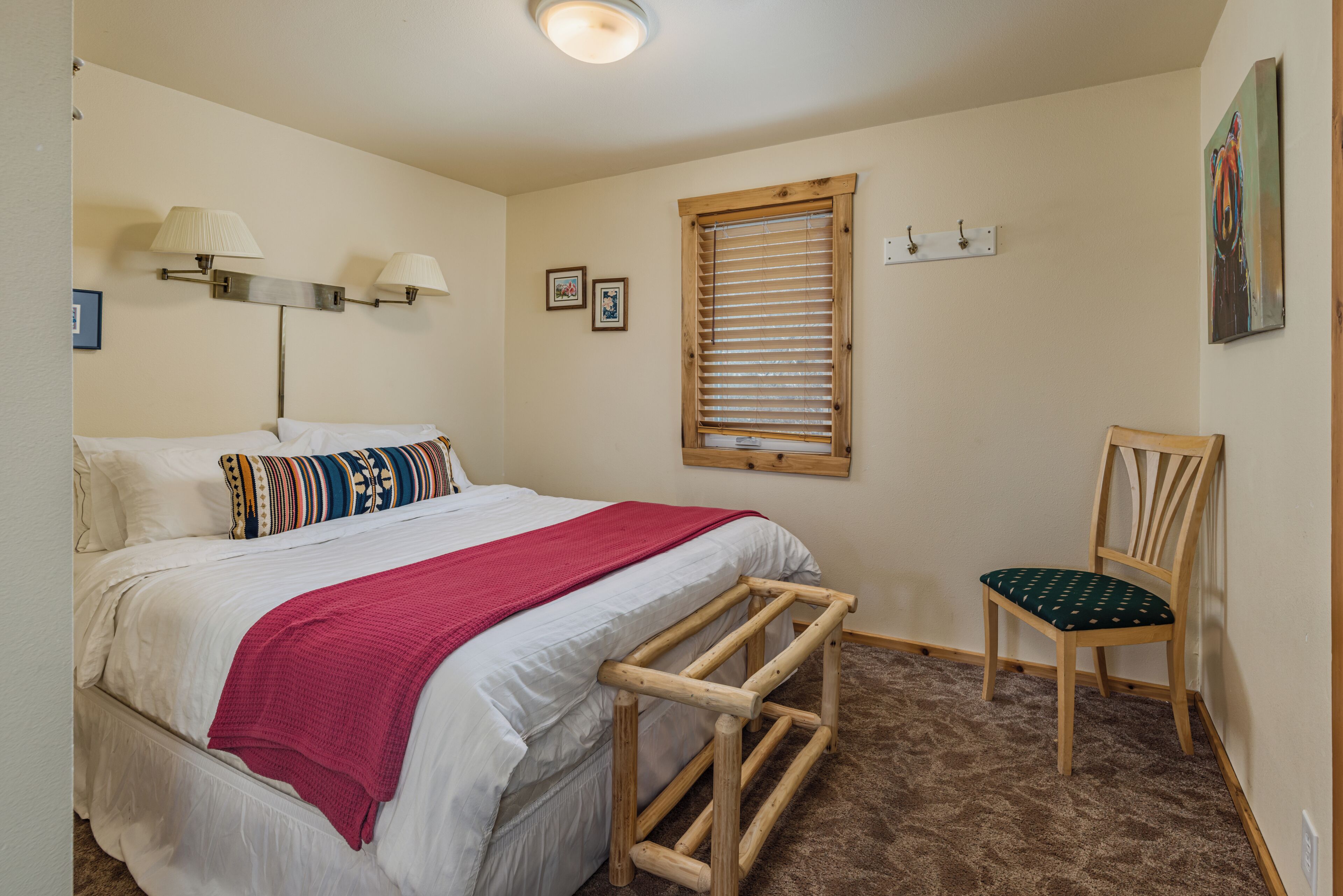 Un dormitorio con cama, una silla, ventana con persianas, lámparas de pared y un cuadro.
