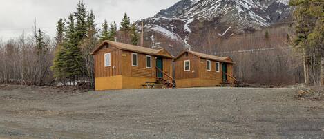 A two-story wooden cabin with a front porch, a staircase, and a view of a snow-capped mountain.